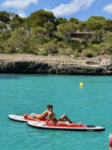 Relaxing women sunbathing on stand-up paddleboard in turquoise sea with lush green trees and modern houses in the background, Sardinia, Italy.