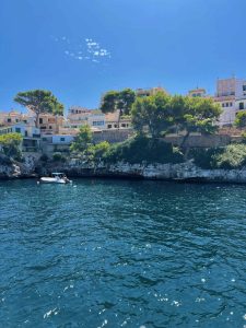 Boats floating in a clear blue sea with a scenic coastal village featuring white buildings and lush trees on a sunny day.